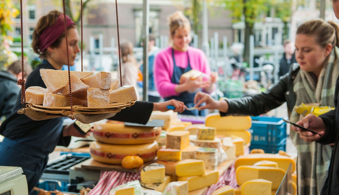 Market scene with cheese samples on a hanging scale and vendors serving customers, with a blurred canal background.